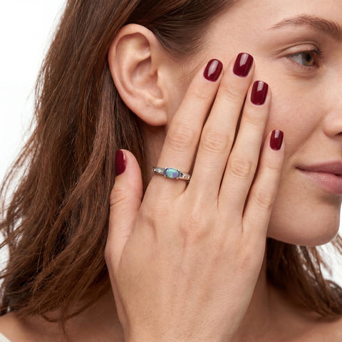 A close-up lifestyle shot of a woman wearing a silver ring with a small oval blue opal gemstone on her finger, with her hand resting against her cheek.