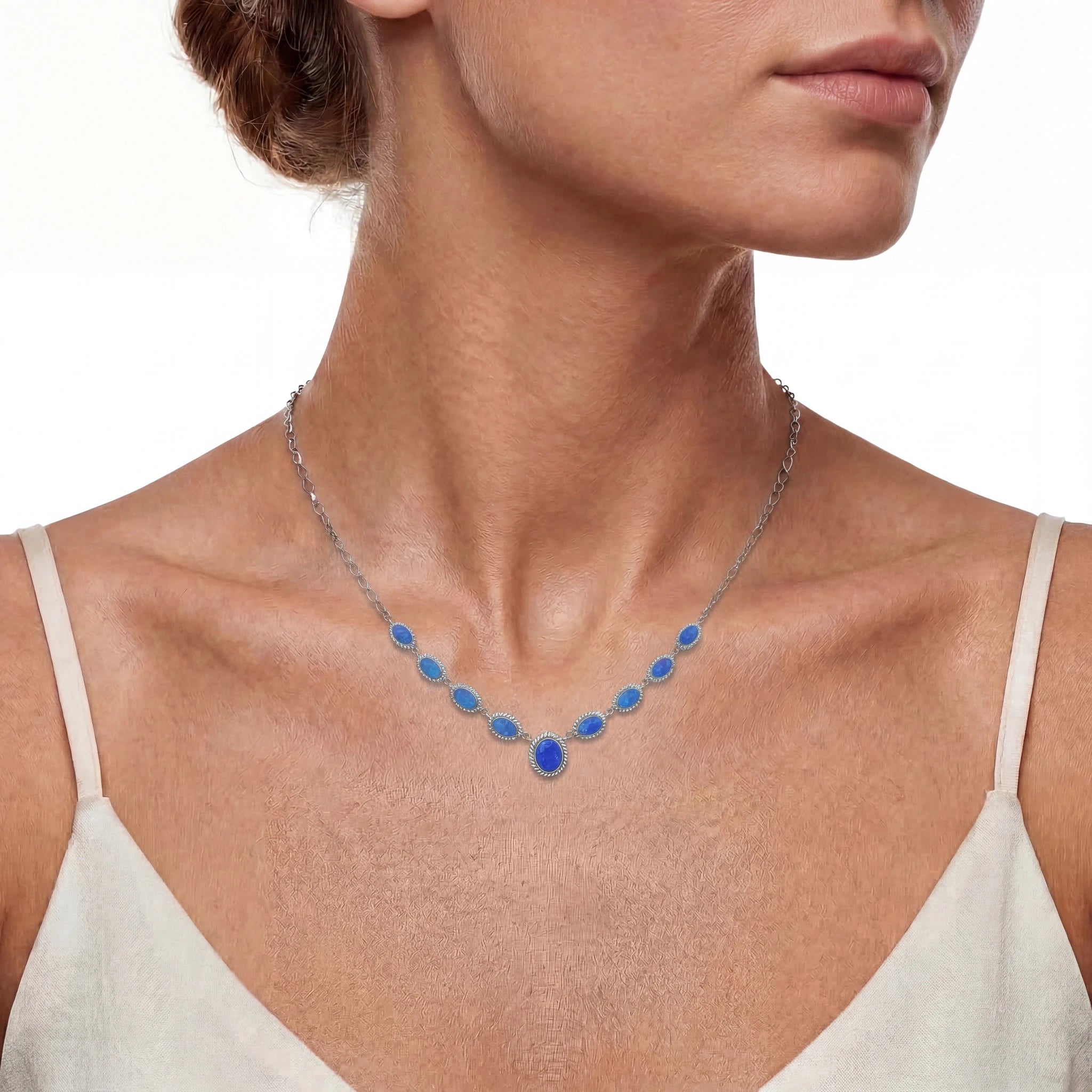 A close-up of a woman wearing a delicate silver necklace with a series of small blue oval opal stones leading to a larger blue teardrop pendant at the centre of the chest.
