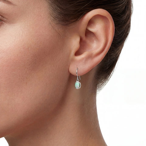 A close-up of a woman's ear wearing a small silver drop earring with a light mint green oval stone, set against a clean white background.