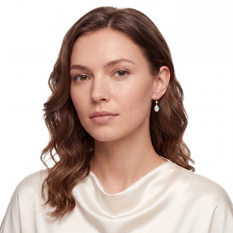 A woman with wavy brown hair wearing a cream satin top and a small silver-toned dangle earring with a white teardrop stone, photographed against a plain white background.