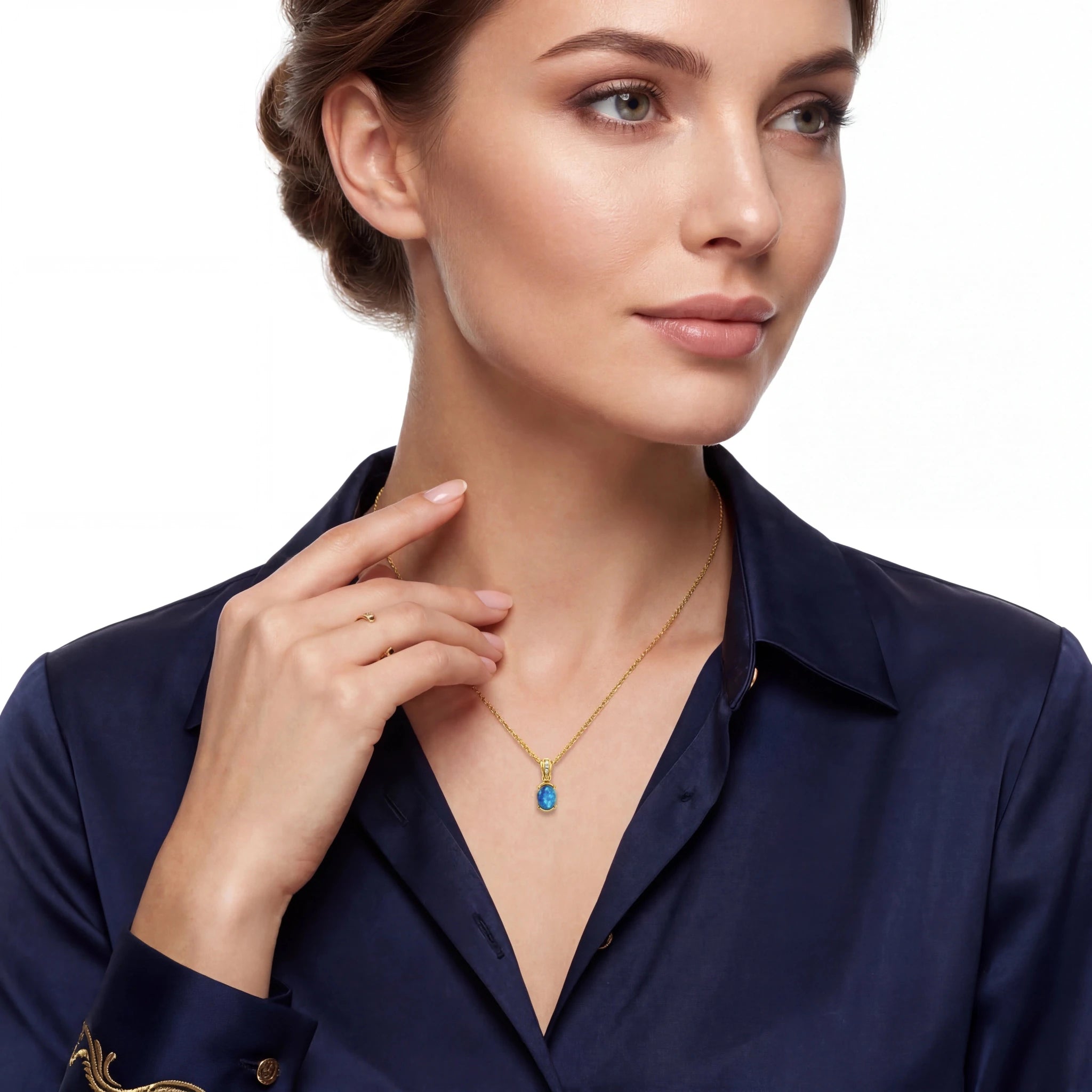 A woman wearing a delicate gold necklace with a small blue teardrop pendant, styled with a navy blue satin shirt against a clean white background.