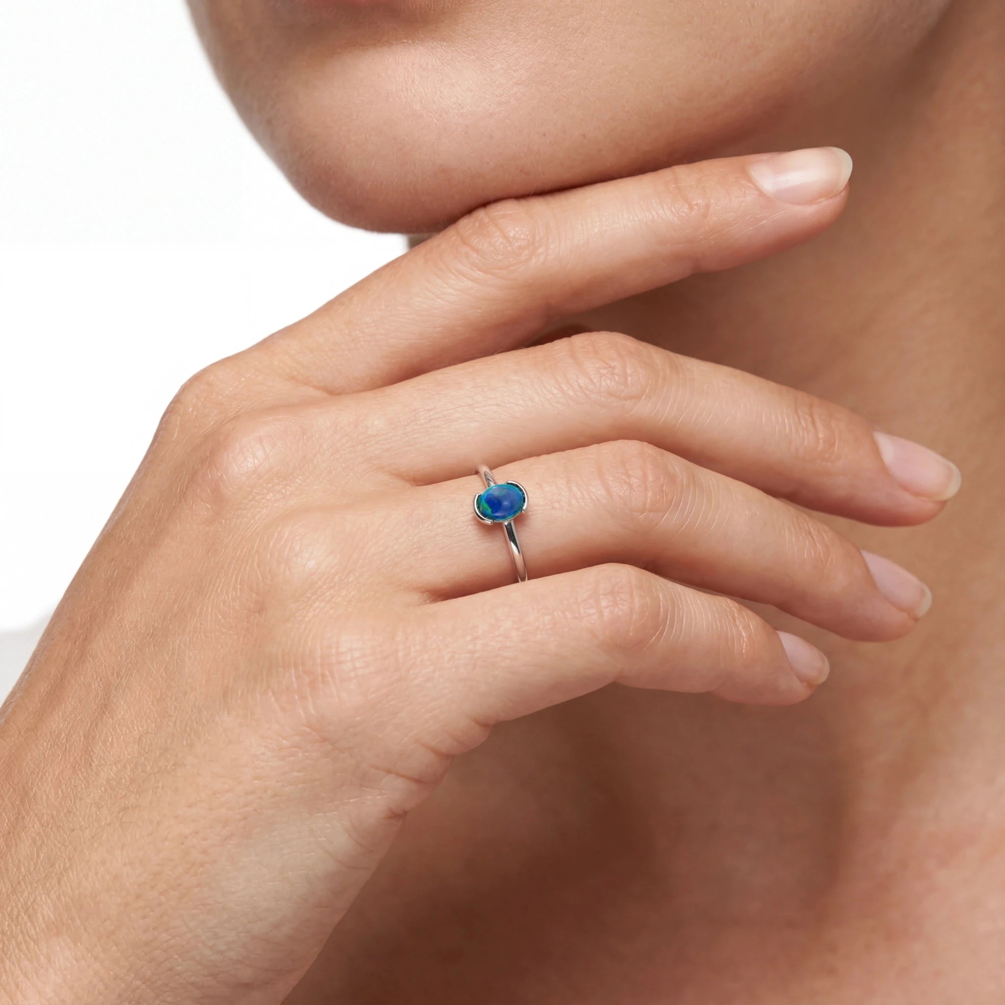 A close-up of a person's hand wearing a silver ring with a small oval blue gemstone, set against a soft white background.