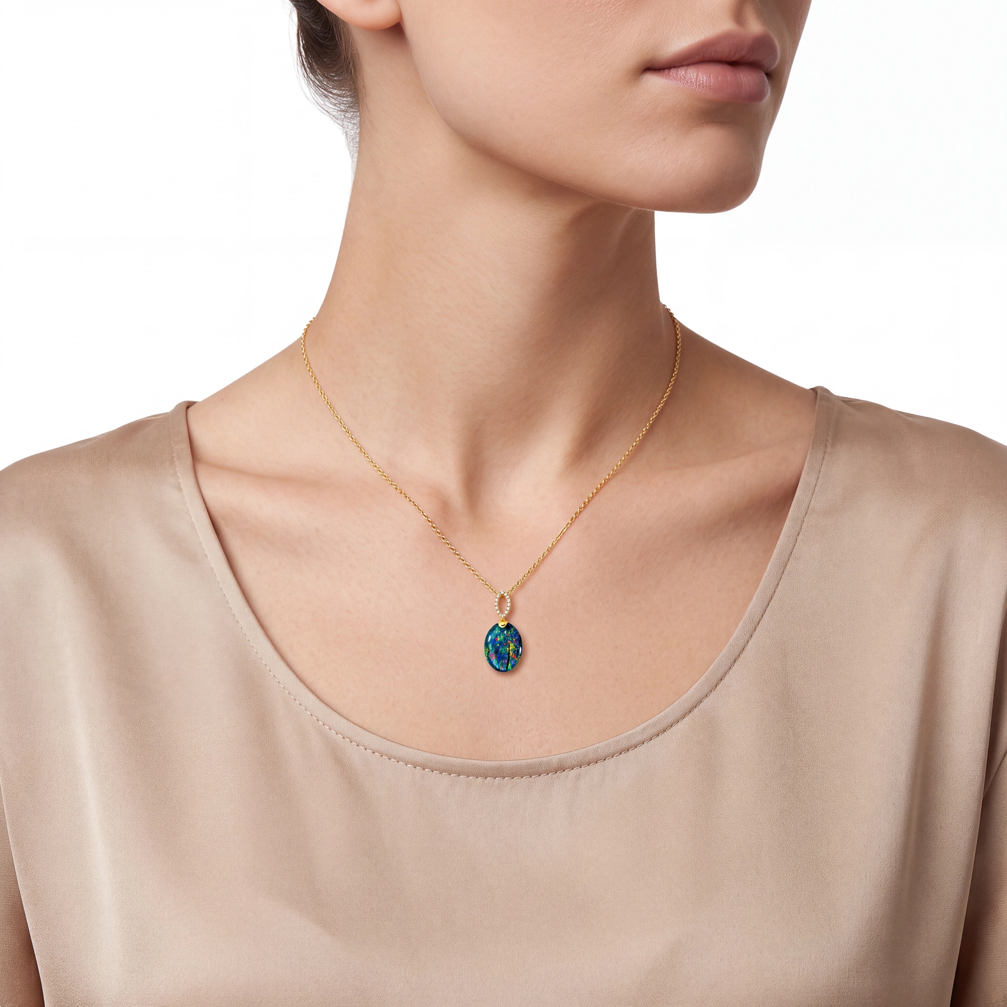A close-up of a model wearing a delicate gold chain necklace with a small oval gold pendant and a blue-green teardrop-shaped stone pendant, paired with a taupe satin top against a white background.