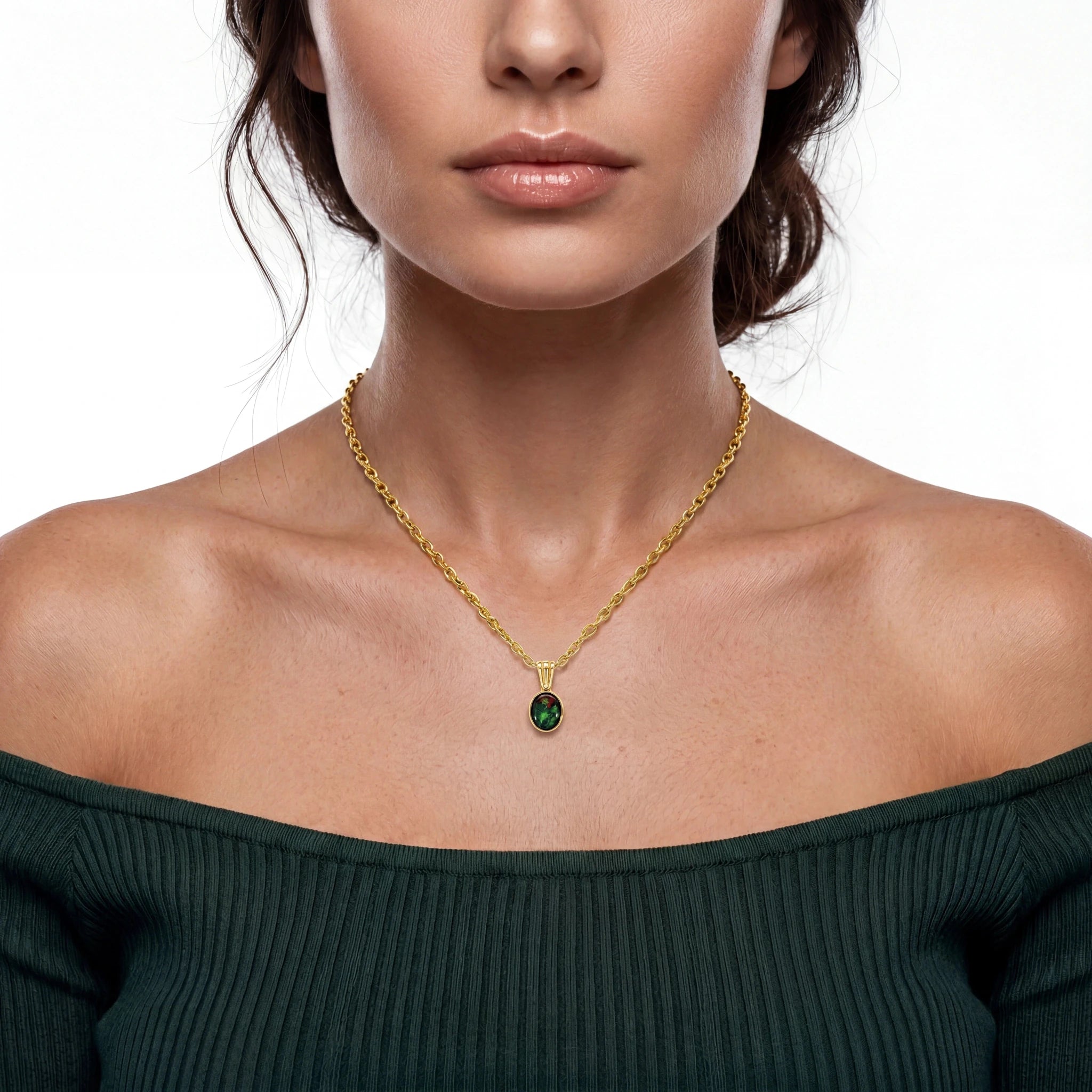 A close-up of a model wearing a delicate gold chain necklace with a small oval green pendant, paired with a dark green off-the-shoulder top against a white background.