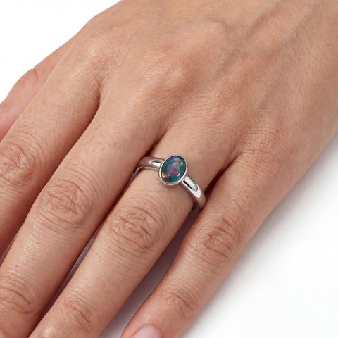 A silver ring with an oval iridescent opal gemstone worn on a person's finger, shown against a clean white background.