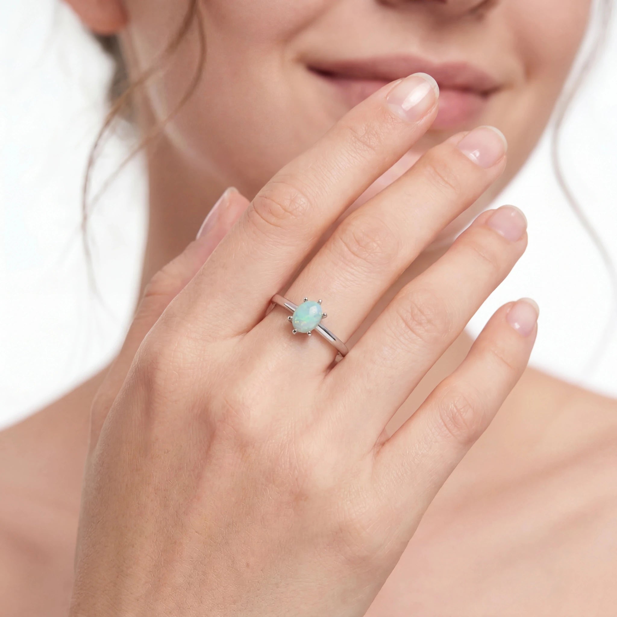 A close-up of a woman's hand wearing a silver ring with a small oval light blue-green gemstone, set against a soft white background.