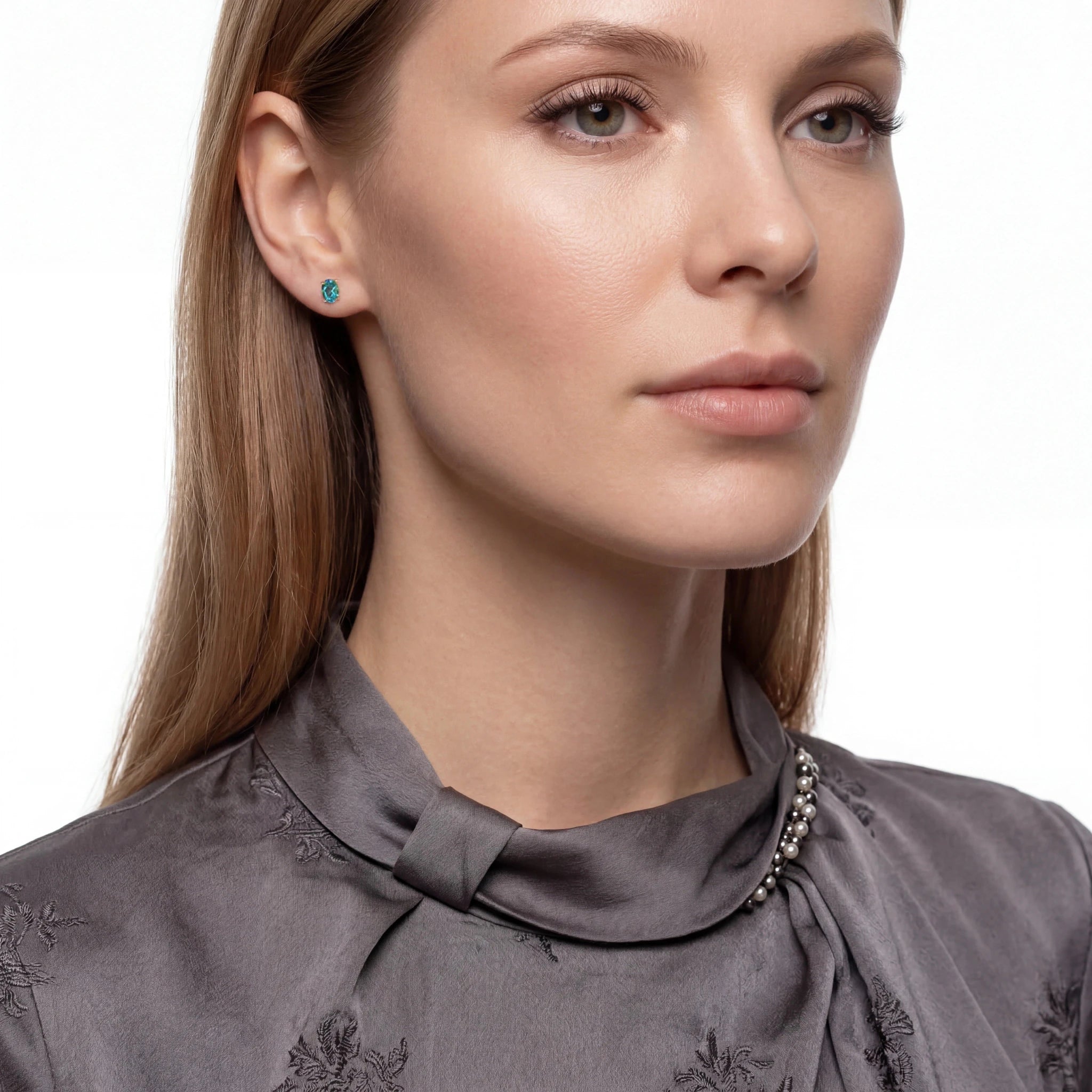 A close-up portrait of a woman wearing a small round turquoise-blue stud earring and a gray satin high-neck blouse with embroidered details.