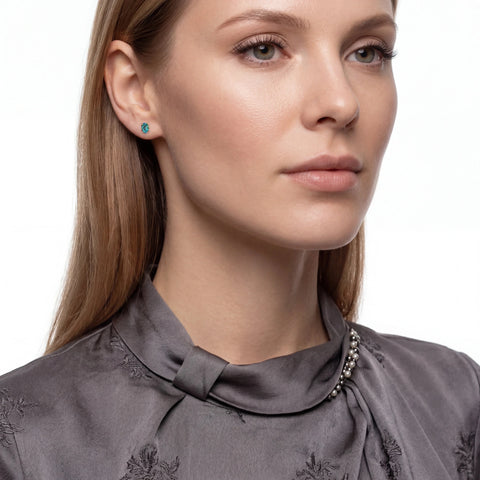 A close-up portrait of a woman wearing a small round turquoise-blue stud earring and a gray satin high-neck blouse with embroidered details.