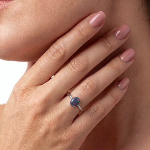 Hand wearing a ring with a blue gemstone, pink nail polish on a neutral background