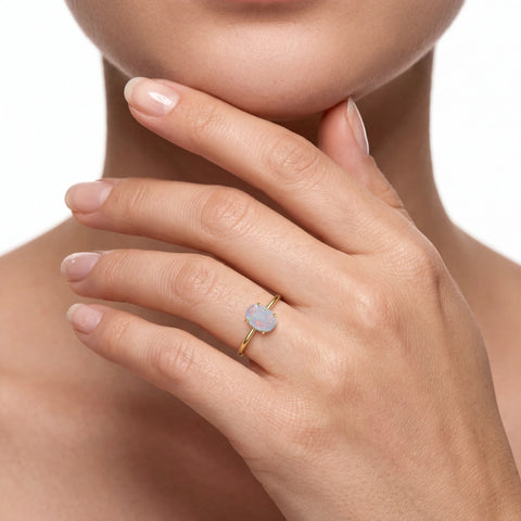 A close-up of a model's hand wearing a delicate gold ring with a small, pale blue-grey opal gemstone.