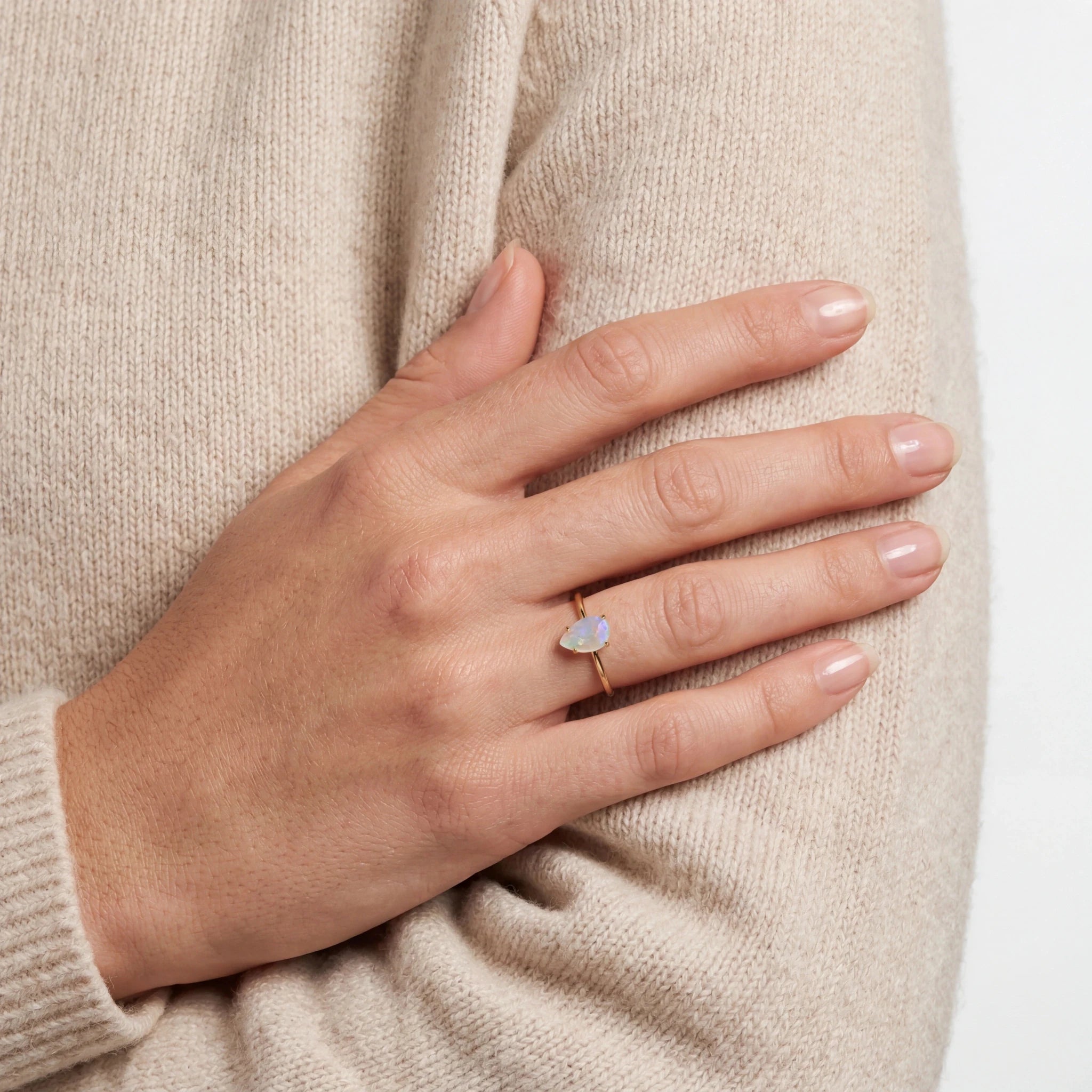 A close-up of a woman's hand resting on their arm, wearing a delicate gold ring with a small iridescent white heart-shaped opal stone.