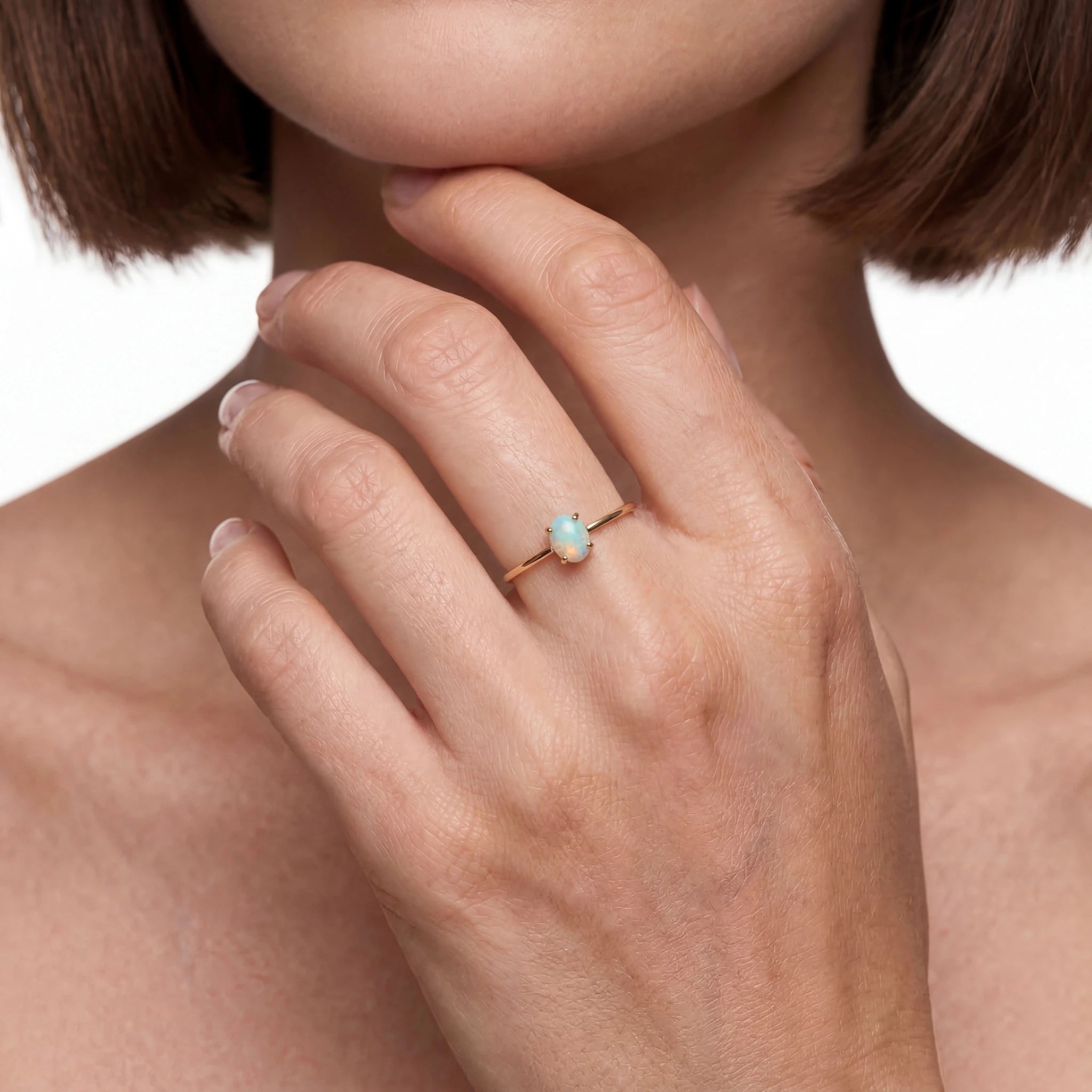 Close-up of a hand wearing a ring with an opal stone, touching the chin against a neutral background.