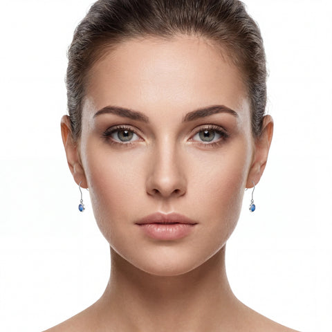 A close-up portrait of a woman with her hair pulled back, wearing small dangling earrings with blue teardrop-shaped stones on silver hooks, against a plain white background.