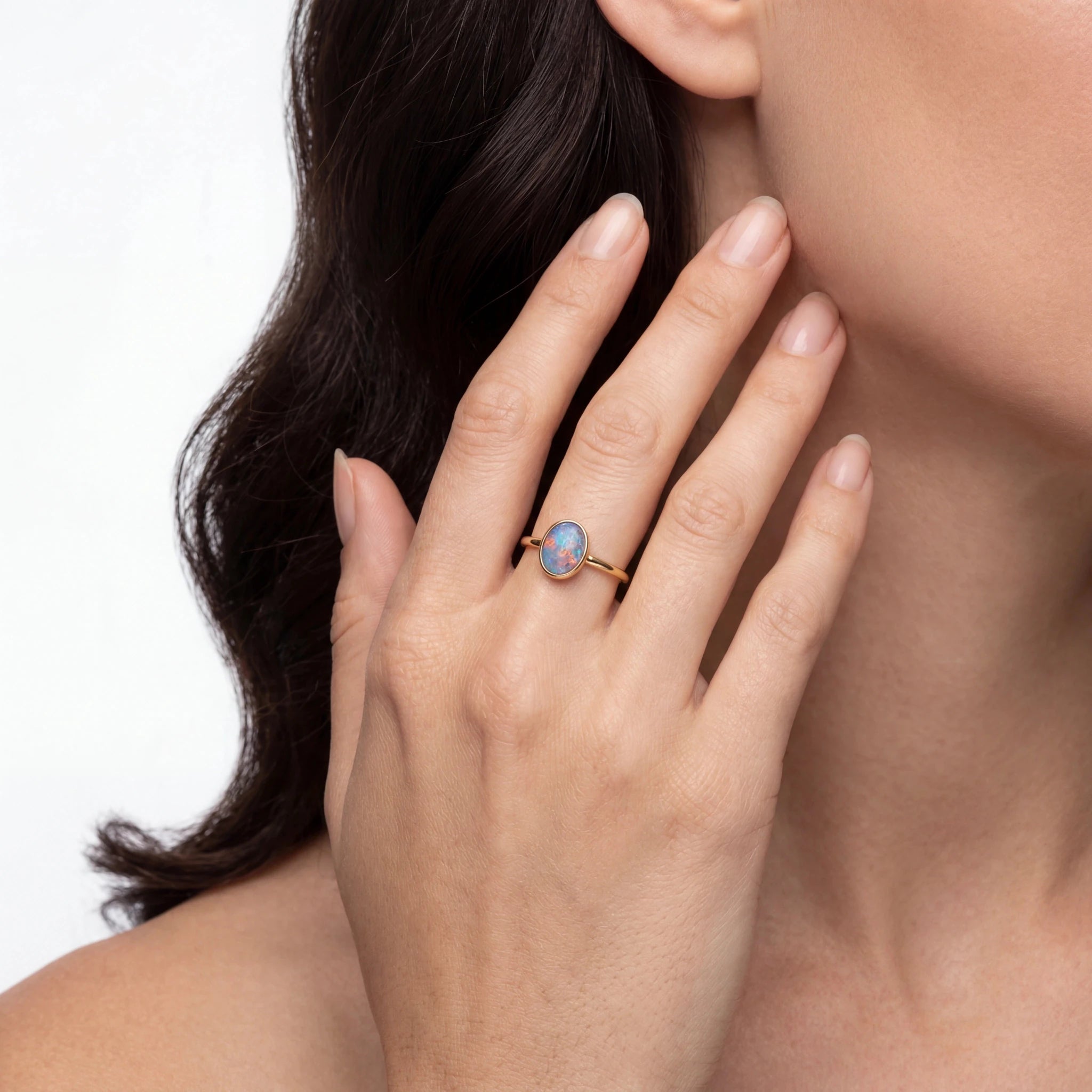 Close-up of a woman wearing a ring with a blue opal gemstone, touching the neck against a neutral background