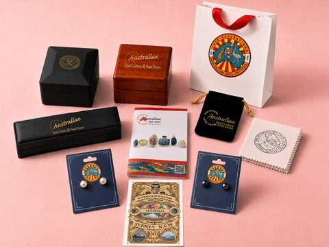A curated flat-lay of Australian-themed gift items on a solid pink background, including jewellery earrings on blue cards, a wooden and black presentation box, a white gift bag, a black velvet pouch, and a small printed booklet.